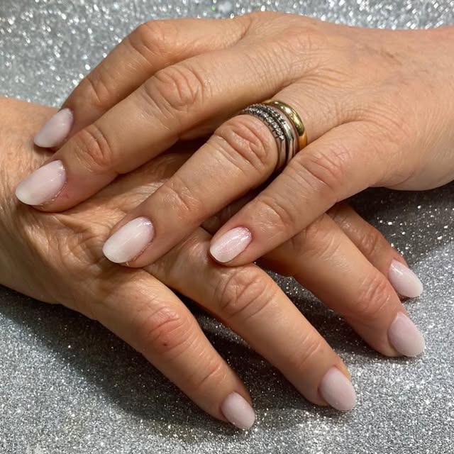 Due mani appoggiate su fondo argento glitterato, unghie ovali smaltate in rosa latte perlato, anelli sottili a fascia.