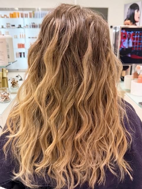 Capelli lunghi biondo caramello visti di spalle in salone, onde morbide su tutta la lunghezza e radici piu scure sfumate.